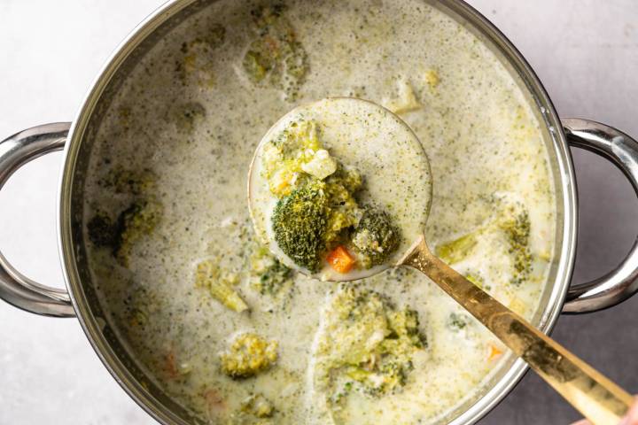 Delicious broccoli cheese soup in a pot with creamy texture, carrots, and broccoli florets. A ladle lifts a hearty portion, conveying warmth.